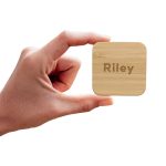 A hand holding a square bamboo wireless charger with the name "Riley" engraved on it.