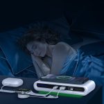 A wireless charging station in white and green, charging devices beside a sleeping person in a dimly lit bedroom.