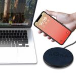 A wireless charger with a circular design in black, positioned next to a laptop and a smartphone.
