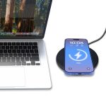 A black wireless charger with a smartphone displaying a charging indicator, beside a laptop with a natural backdrop.