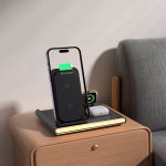 A magnetic wireless charger in black, positioned next to a phone on a wooden bedside table, with a small notebook and earbuds.