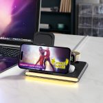 A wireless charger in black with a smartphone, earbuds, and a USB adaptor, set on a desk beside a laptop.