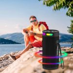 A black wireless speaker with colourful LED lights, positioned by a lake with a person in the background.