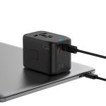 A compact black travel adaptor with multiple ports sits on a laptop, featuring a logo and connected cables.