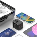 A black travel adaptor with multiple ports, placed beside a smartphone, passport, and a clear case on a flat surface.