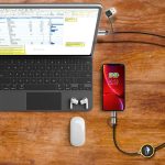 A LED charging cable in black, connected to a smartphone and a laptop, on a wooden table with accessories.
