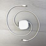 A retractable charging data cable in white, coiled with connectors at both ends, laid on a light wooden surface.