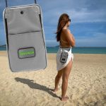 A grey eco-friendly bag displayed on a beach, featuring a logo, with a person wearing it while facing the ocean.