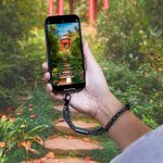 A smartphone is held by a hand with a black and white polka dot wrist strap, set against a lush garden background.