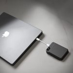 A black magnetic wireless power bank connected to a laptop on a light surface. The power bank has a logo.