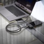 A grey and black charging cable connected to a smartphone and a laptop on a glass table.