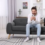 A man seated on a grey couch using a phone, with a charging cable visible on the floor. The setting is bright and modern.