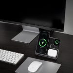 A wireless charging stand in black with compartments for a phone, AirPods, and a smartwatch, placed on a desk.