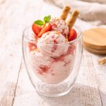 A glass dessert cup filled with pink ice cream, topped with strawberries and two wafer sticks, garnished with mint leaves.