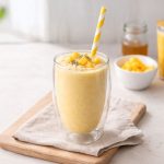 A drink bottle featuring a clear double wall design filled with a yellow smoothie topped with mango and chia seeds, with a striped straw.