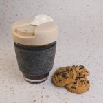 A reusable coffee cup in grey with a lid, featuring a felt band, accompanied by chocolate chip cookies.