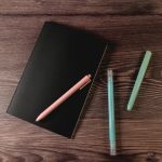 A black notebook placed on a wooden surface, accompanied by a pink pen and two teal-colored pens.
