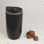 A reusable coffee cup with a sleek black design, featuring a secure lid, accompanied by several chocolate coffee pods.