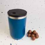 A blue reusable coffee cup with a black lid and a silver rim, accompanied by several brown chocolate balls.
