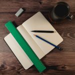 A green 30cm recycled plastic ruler rests on an open dotted notebook with a black pen, blue pencil, and a coffee cup.