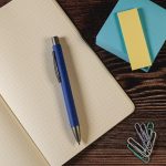 A blue Aspen Paper Pen rests on a dotted notebook, accompanied by sticky notes and paper clips.