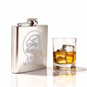 A stainless steel hip flask sits beside a glass of whiskey with ice, featuring an engraved design and a logo.