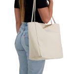 A cream-coloured tote bag is held by a person wearing a black top and blue jeans. The bag has a simple design and a logo.