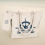 A canvas tote bag in cream with a nautical design and rope handles, featuring a matching tag.