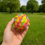 A hand holds a vibrant, multicoloured rubber ball with textured bumps, set against a green park background.