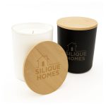 A medium-sized glass candle with a white container, wooden lid, and a black container beside it, both featuring a logo.