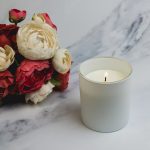 A medium glass candle in a white holder beside a bouquet of red and white roses on a marble surface.