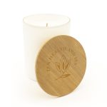 A large white glass candle with a wooden lid, featuring a logo and an embossed design on the lid.