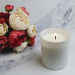 A large white glass candle with a lit wick beside a bouquet of cream and pink flowers.