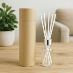A glass reed diffuser with white reeds, in a cylindrical cardboard container, next to a potted plant.