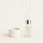 A vanilla reed diffuser with white reeds, housed in a glass bottle with a silver collar, alongside a white container.