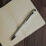 A metal pen in white and black rests atop an open lined notebook on a wooden surface. The pen features a logo.
