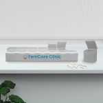 A white weekly pill box with multiple compartments, some open, alongside white pills and a plant. It has a logo.