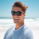 Bamboo sunglasses with dark lenses, worn by a smiling man in a striped shirt, set against a beach backdrop.