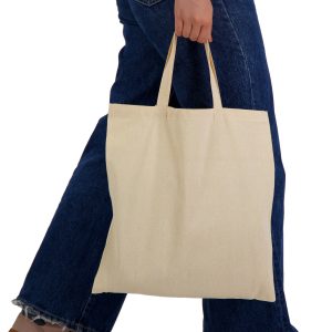 A tote bag in a natural cream colour with short handles, being held by a person wearing blue denim jeans.