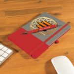 A grey notebook with a red band and a matching red pen placed on a wooden desk alongside a keyboard and mouse.