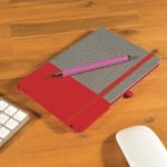 A Venture Bondi Notebook with a red and gray cover is featured alongside a pink Austin Pen on a wooden surface.