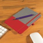 A grey notebook with a red section alongside a purple pen, placed on a wooden desk next to a keyboard and mouse.