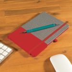 A notebook with a gray cover and a red bottom, paired with a teal pen, on a wooden desk.