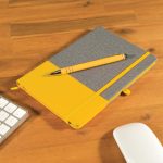 A Venture Bondi Notebook in gray and yellow with an Austin Pen resting on top, placed on a wooden desk.