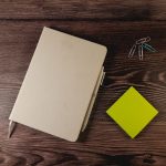 An A5 notebook in a beige cover with a pen, alongside colourful paper clips and a green sticky note on a wooden surface.