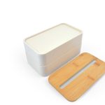 A two-tiered lunch box in cream color with a bamboo lid designed to hold a phone. It features a simple logo.