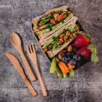 A lunch box filled with wholemeal wraps, strawberries, blueberries, celery sticks, and almonds, accompanied by wooden utensils.