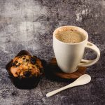 A cream-colored reusable coffee cup with a wooden base, spoon, and a chocolate chip muffin. The cup features a logo.