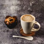 A white wheat fibre cup with a spoon, resting on a wooden coaster beside a chocolate chip muffin.