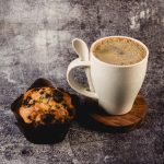 A white wheat fibre cup with a spoon, filled with coffee, alongside a chocolate chip muffin on a wooden coaster.